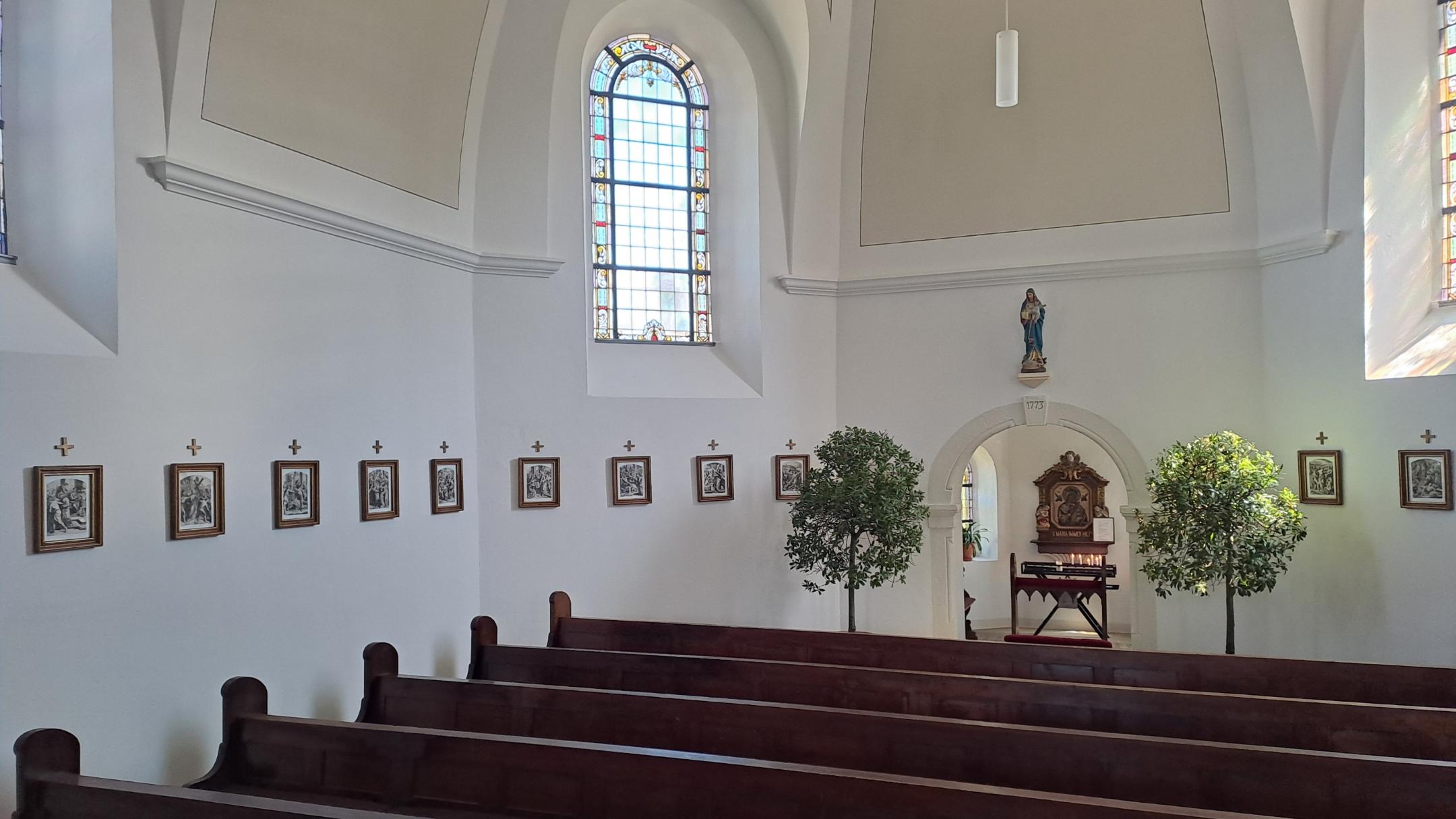 Seitenschiff mit mehreren Holzbänken und einer Reihe kleiner gerahmter Kreuzwegstationen an der weißen Wand. Am Ende des Raums steht eine kleine Kapelle mit Marienbild.