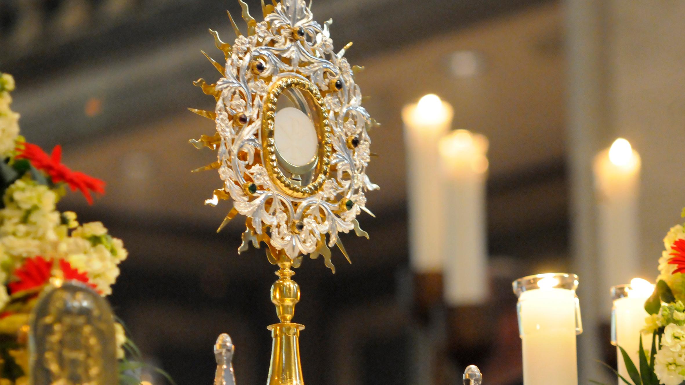 Eine reich verzierte Monstranz steht auf einem Altar in einer Kirche. In der Mitte der Monstranz ist die weiße Hostie sichtbar. Die Monstranz besteht aus gold- und silberfarbenem Metall mit strahlenförmigen Ornamenten. Rundherum stehen mehrere brennende Kerzen in Glasgefäßen. Blumenschmuck rahmt den Altar. Der Hintergrund ist unscharf und zeigt das Innere des Kirchenraums.